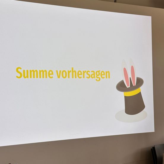 Ein Whiteboard mit den Worten 'Summe vorhersagen' in gelber Schrift und einem Zauberhut mit Hasenohren in der unteren rechten Ecke.