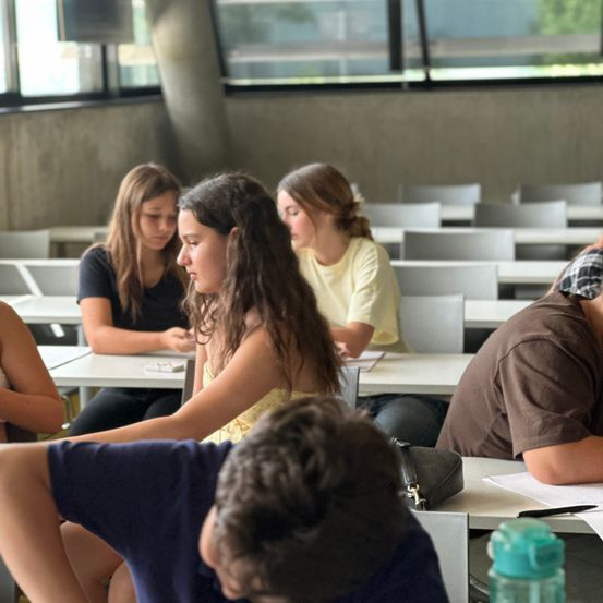 Ein Klassenzimmer mit Schülern, die an Tischen sitzen und sich auf ihr Studium konzentrieren. Einige schauen auf Papiere, während andere in ein Gespräch vertieft zu sein scheinen. Der Raum hat Glasfenster und Betonwände.