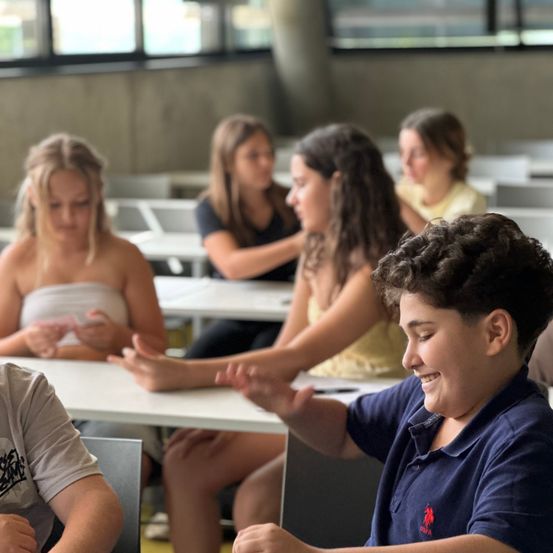 Mehrere Personen sitzen in einem Klassenzimmer. Ein Mädchen im weißen Tanktop spricht mit einem anderen Mädchen mit lockigen Haaren. Ein Junge im blauen Poloshirt lächelt und macht eine Geste.