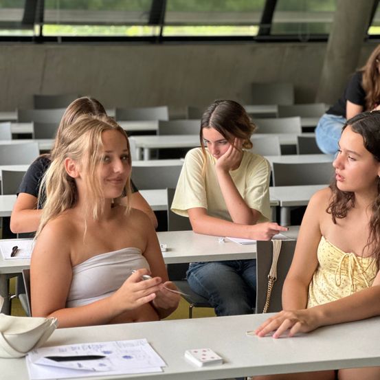 Eine Gruppe von Mädchen in einem Klassenzimmer, ein Mädchen lächelt und hält etwas, ein anderes schaut sie an und ein drittes hat die Hand im Gesicht. Papiere und ein Stift auf dem Schreibtisch.