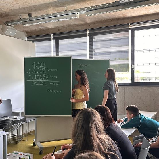 Zwei Frauen stehen vor einer Tafel mit Gleichungen und Zahlen und unterrichten einen Klassenraum. Die Schüler sitzen aufmerksam, und ein Junge vorne sieht engagiert aus.
