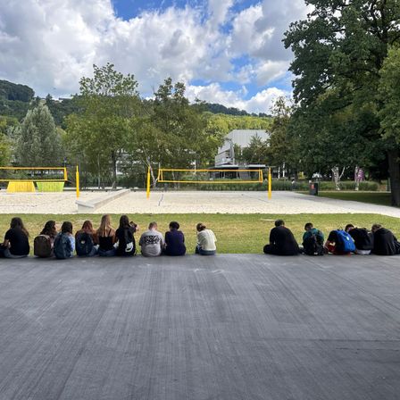 Eine Gruppe von Menschen sitzt auf dem Betonboden, vielleicht um ein Spiel zu beobachten. Einige von ihnen tragen Rucksäcke. Vor ihnen stehen zwei Volleyballnetze. Das Gebiet ist von Bäumen und Gras umgeben. In der Ferne befindet sich ein Gebäude. Der Himmel ist blau mit Wolken.
