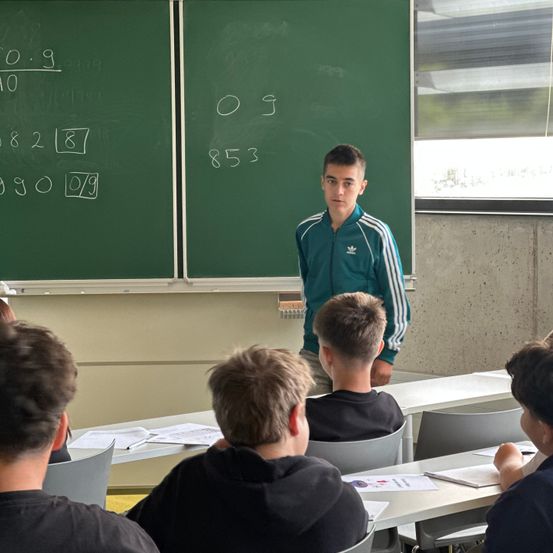 Ein junger Mann steht vor einem Klassenzimmer und spricht Schüler an, die an Tischen sitzen. Hinter ihm zeigt eine grüne Tafel mathematische Gleichungen.