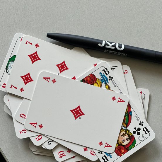 Ein Stapel Spielkarten mit einem Stift, der mit JKU beschriftet ist, auf einer grauen Oberfläche. Die Karten beinhalten Gesichter wie den Joker und den Ass der Pik.