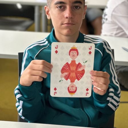 Ein junger Mann hält eine Spielkarte mit einer roten Jack-Figur in der Hand, die eine Krone trägt und eine Blume hält. Er ist in einem Klassenzimmer.