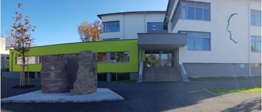 Ein weißes und grünes Gebäude mit Glasfenstern und einem Treppeneingang. Zwei große Steine stehen vor dem Gebäude. Ein Baum befindet sich auf der linken Seite. Der Himmel ist klar.
