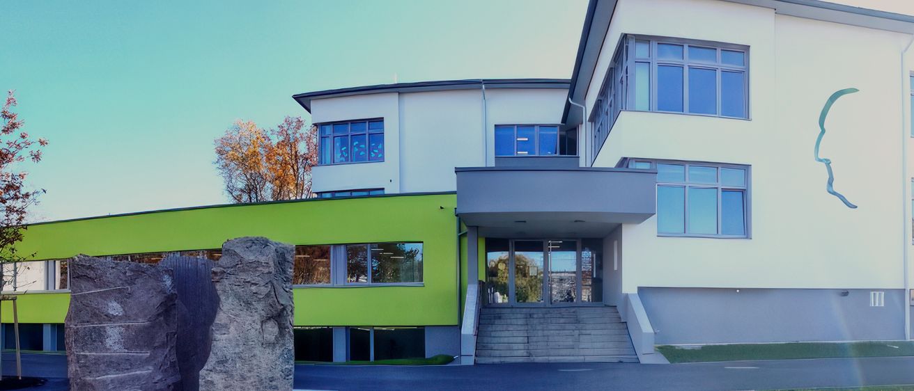 Ein modernes Gebäude mit einer grünen Fassade und zwei großen Steinskulpturen davor. Das Gebäude hat Glasfenster und eine Treppe, die zum Eingang führt. Der Bereich ist von einer Betonauffahrt umgeben.