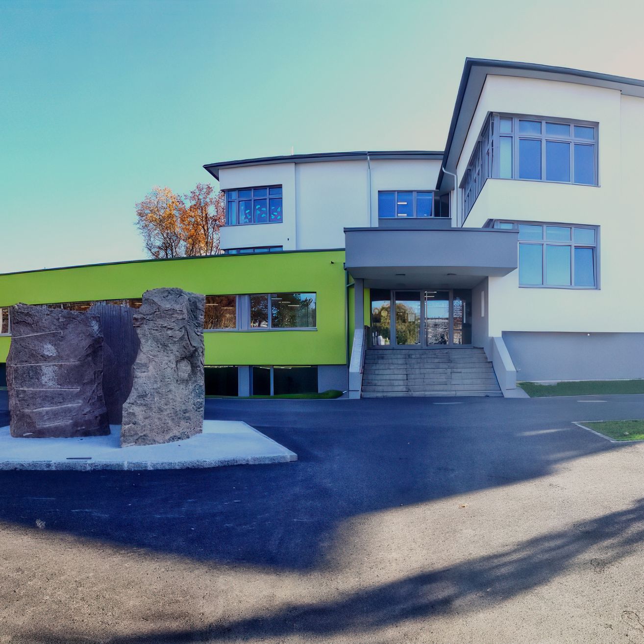 Ein modernes Gebäude mit Glasfenstern und einer grünen Akzentwand steht vor einem klaren Himmel. Zwei große Steine sind vor dem Eingang platziert.