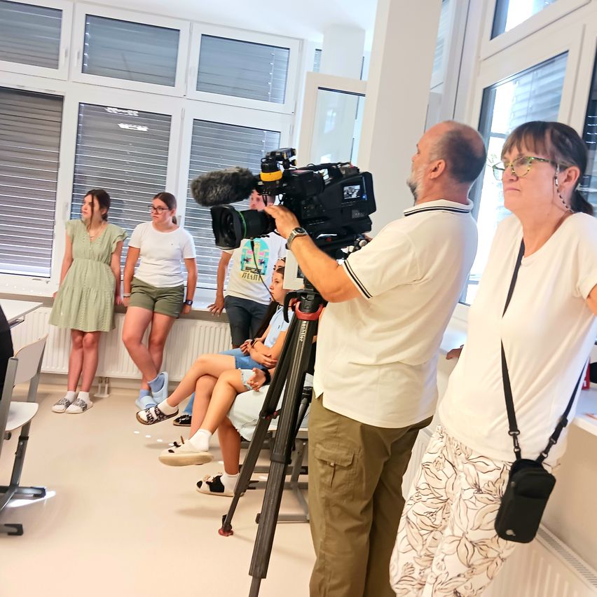 Ein Mann mit einer Kamera filmt in einem Raum. Zwei Frauen beobachten. Mehrere Personen sitzen auf einer Bank. Der Raum hat weiße Wände und große Fenster.