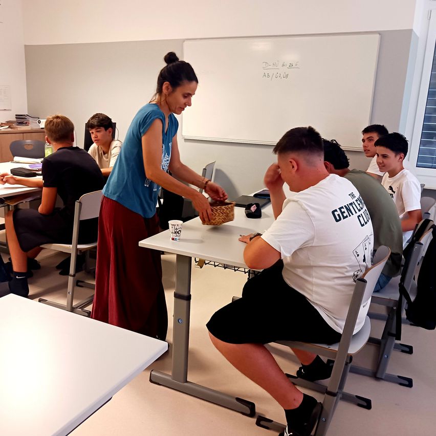 Eine Frau steht an einem Schreibtisch und verteilt Gegenstände an Schüler, die an Tischen in einem Klassenzimmer sitzen. Die Schüler tragen Freizeitkleidung, und der Raum hat weiße Wände und Tische.