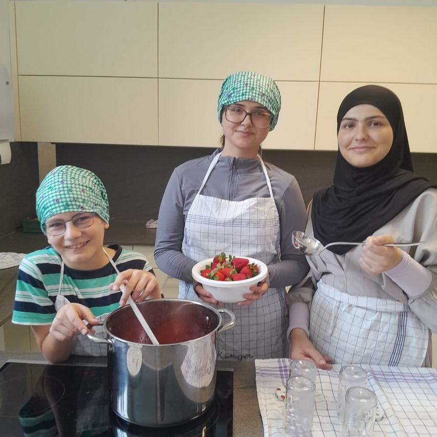 Drei Frauen in Schürzen kochen in einer Küche, eine mit einer Schüssel Erdbeeren, eine andere mit einem Topf und die dritte mit einem Löffel. Dahinter befindet sich eine Theke mit Gläsern und einem Herd.