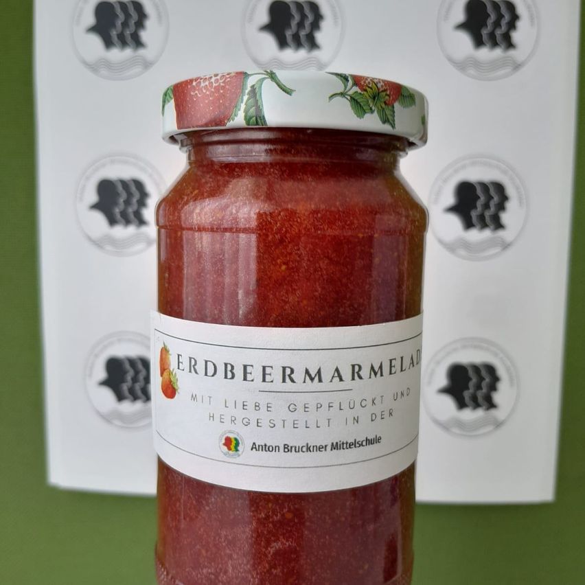 Ein Glas Erdbeermarmelade mit weißem Deckel, liebevoll hergestellt. Das Etikett trägt den Namen 'Erdbeermarmelade' und es wurde in der Anton Bruckner Mittelschule produziert.