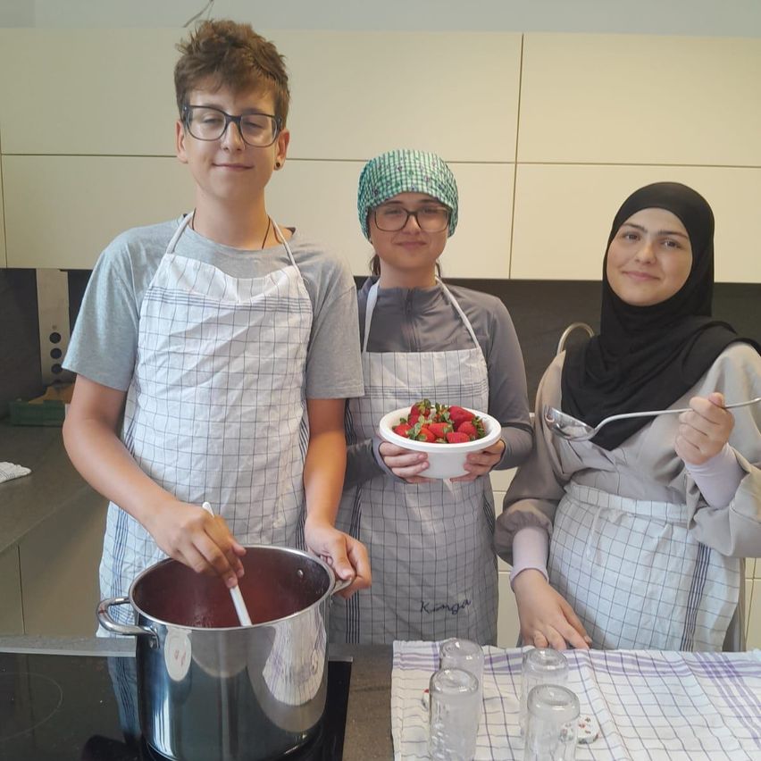 Drei Personen in Schürzen, eine rührt einen Topf, eine andere hält eine Schüssel mit Erdbeeren und die dritte hat einen Löffel, in einer Küche.