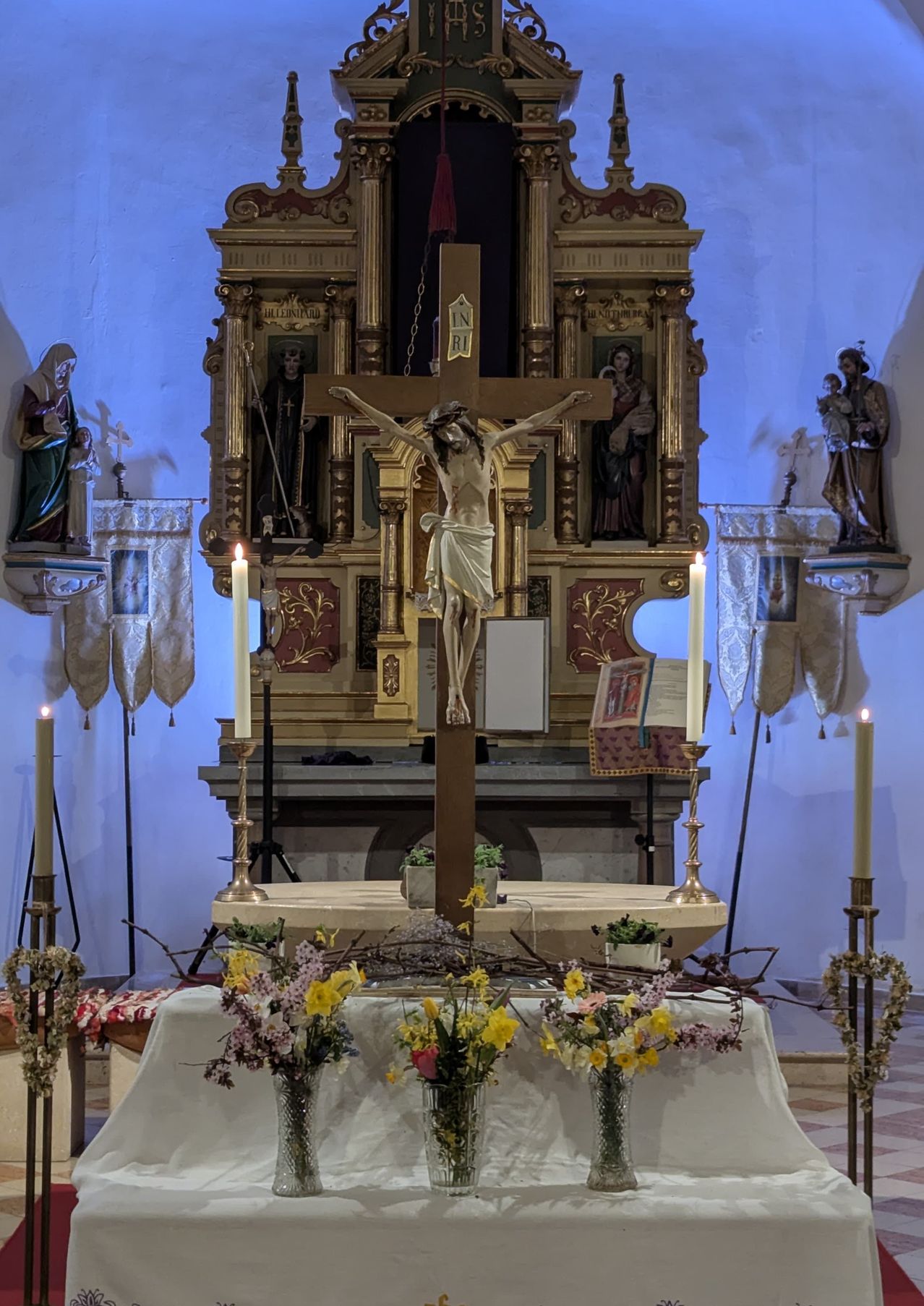 Ein Kirchenaltar mit Kreuz und Jesus-Statue. Vier Kerzen umgeben den Altar, mit Blumen und Kränzen in der Nähe. Über dem Altar sind Heilige-Statuen ausgestellt.