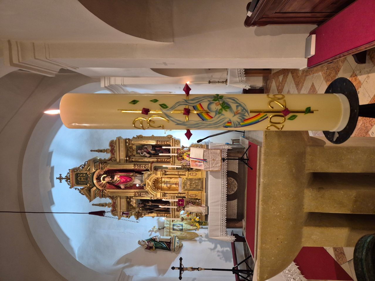 Vor einem Altar mit einer Heiligenstatue steht eine verzierte Kerze. Die Kerze hat ein Regenbogendesign und die Nummer 2020. Hinter dem Altar bedeckt ein weißer Stoff einen Tisch.