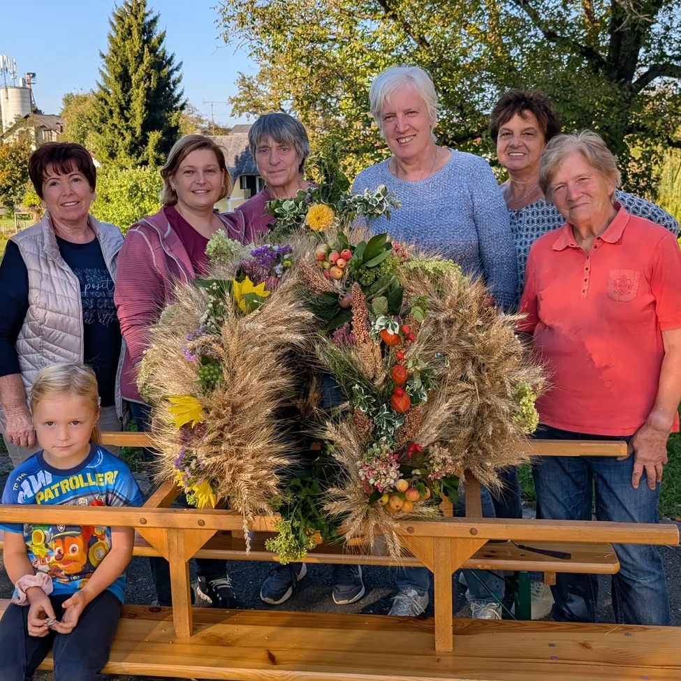 Eine Gruppe von Frauen und einem kleinen Mädchen stehen hinter einer Holzbank mit einer großen Blumendekoration in der Mitte.