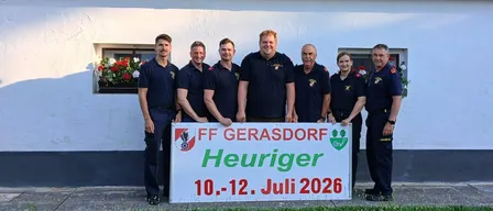 Eine Gruppe von Menschen steht vor einem weißen Gebäude. Sie halten ein Schild mit 'FF Gerasdorf Heuriger 10.-12. Juli 2026'. Sie tragen Uniformen.