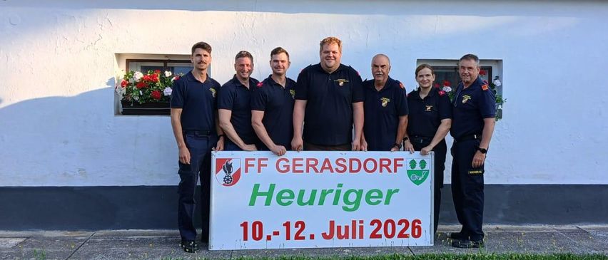 Eine Gruppe von Menschen steht vor einem weißen Gebäude. Sie halten ein Schild mit 'FF Gerasdorf Heuriger 10.-12. Juli 2026'. Sie tragen Uniformen.