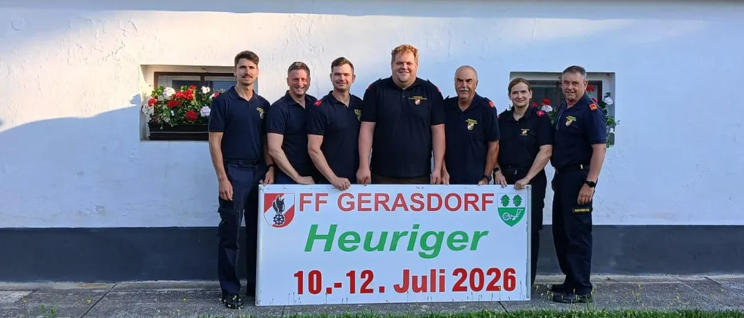 Eine Gruppe von Menschen steht vor einem weißen Gebäude. Sie halten ein Schild mit 'FF Gerasdorf Heuriger 10.-12. Juli 2026'. Sie tragen Uniformen.