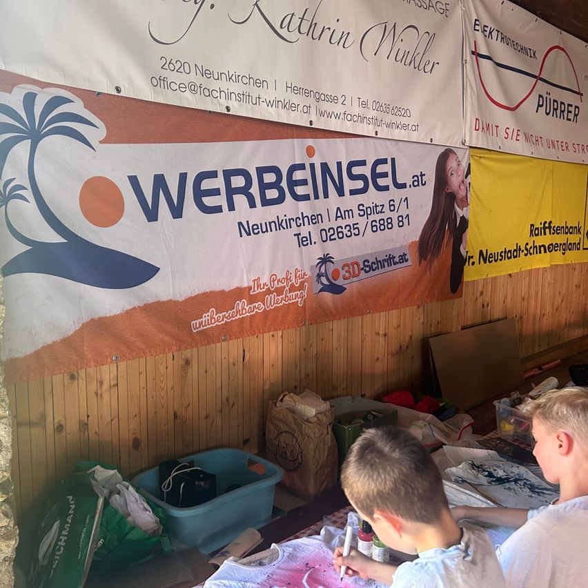 Zwei Jungen bemalen ein weißes T-Shirt mit leuchtenden Farben. Dahinter befindet sich eine Holzwand mit mehreren Bannern, darunter eines mit der Aufschrift 'WERBEISEL.at' und ein weiteres mit dem Bild eines Mädchens.