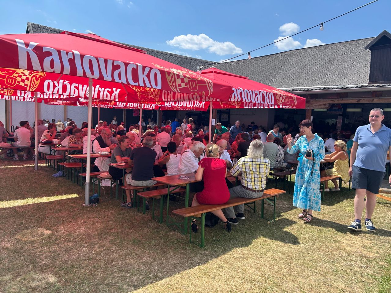 Eine Gruppe von Menschen sitzt an Tischen unter roten Sonnenschirmen. Eine Frau in einem blauen Kleid geht zwischen den Tischen hindurch. Die Umgebung scheint ein Outdoor-Event zu sein, möglicherweise ein Festival.