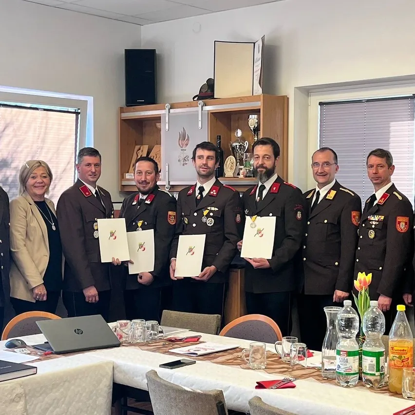 Eine Gruppe von Männern und Frauen in Uniform steht hinter einem Tisch mit Auszeichnungen und Zertifikaten, mit einem Laptop, Flaschen und einer Vase mit Blumen auf dem Tisch. Dahinter steht ein Regal mit Trophäen.