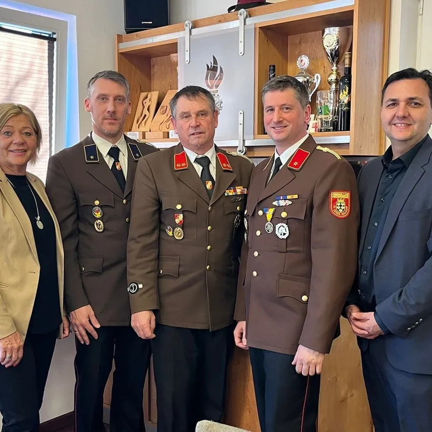 Fünf Personen stehen gemeinsam lächelnd für ein Foto in einem Raum. Drei Männer tragen Militäruniformen mit Medaillen. Eine Frau in einem Blazer steht links. Dahinter zeigt ein Holzschrank Trophäen.