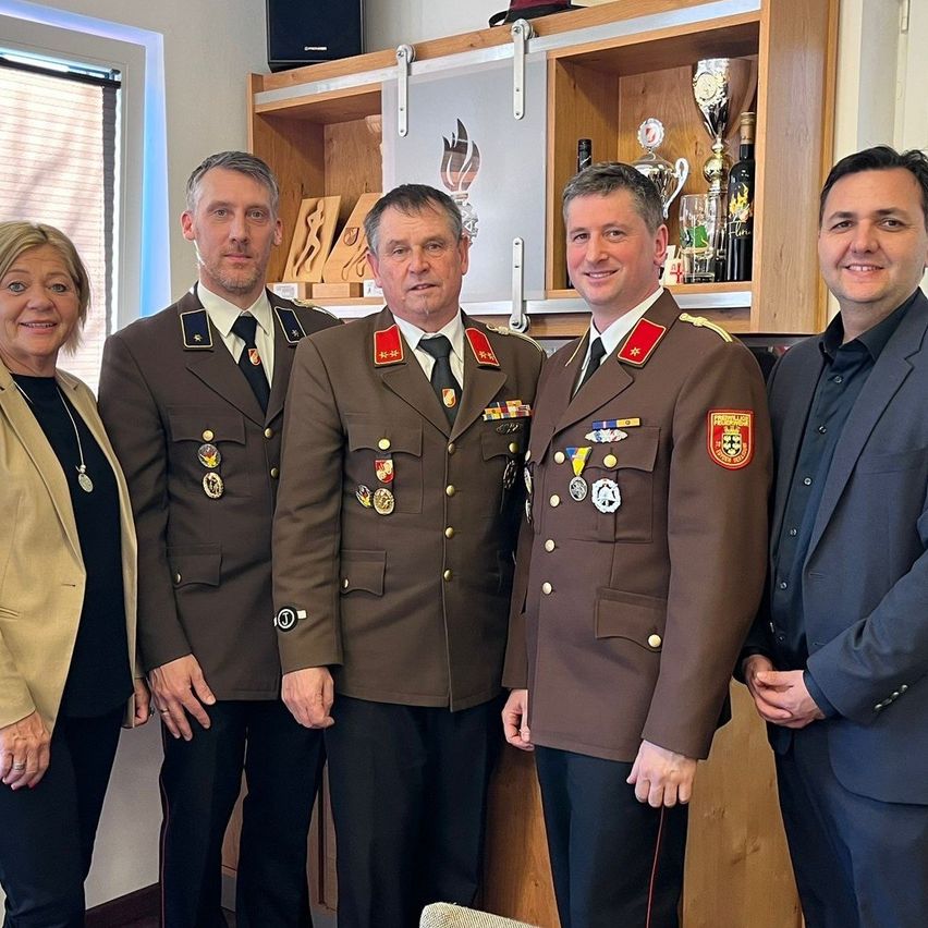 Five individuals stand together, smiling for a photo in a room. Three men wear military uniforms with medals. A woman in a blazer stands on the left. Behind them, a wooden cabinet displays trophies.