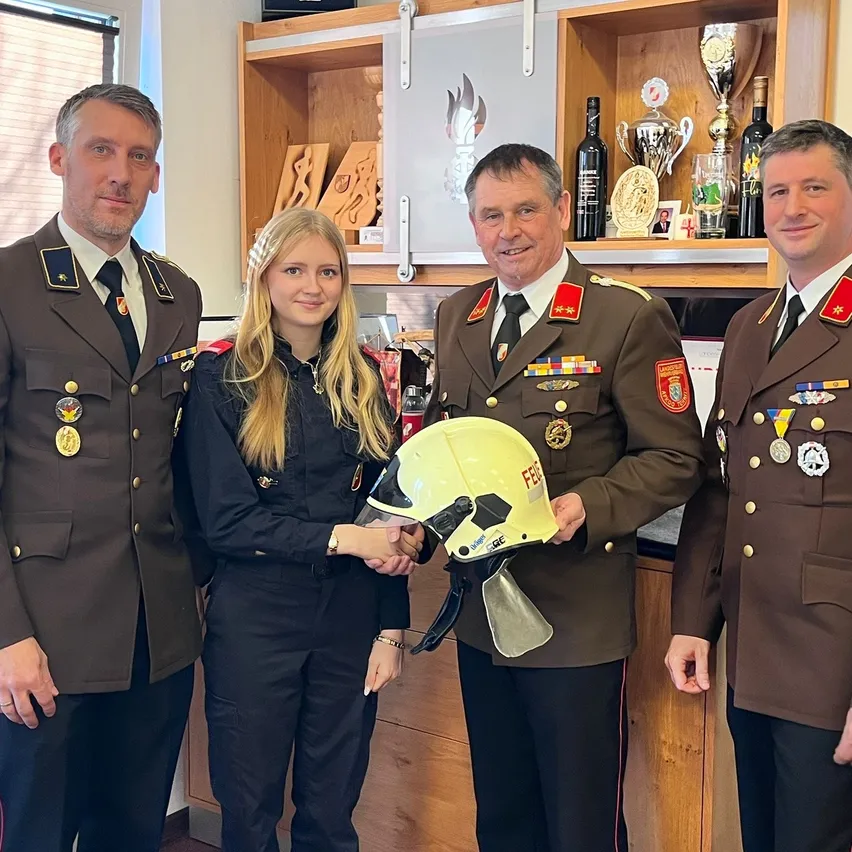 Vier Personen, drei Männer und eine Frau, stehen in einem Raum mit einem Regal dahinter. Die Frau hält einen gelben Helm, und die Männer tragen Militäruniformen und Medaillen.