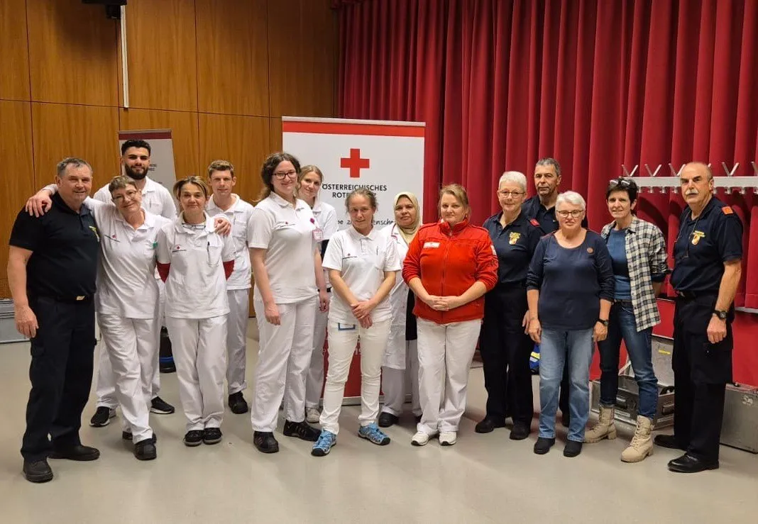 Eine Gruppe von Menschen posiert für ein Foto vor einem roten Banner mit einem weißen Kreuz, möglicherweise für das Österreichische Rote Kreuz.