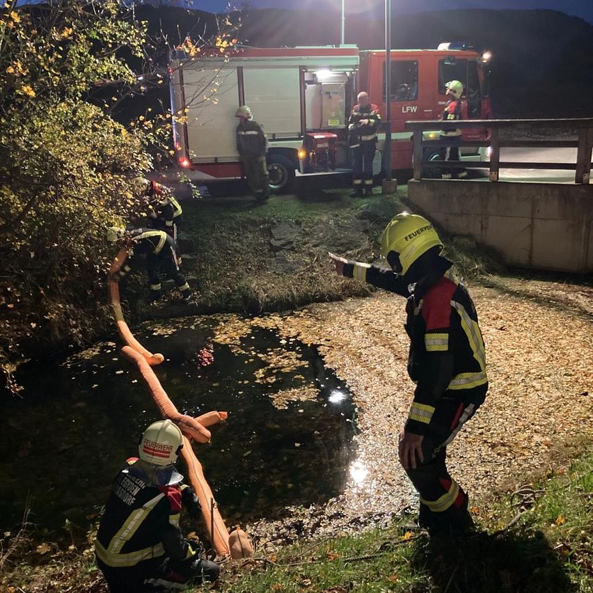 Feuerwehrleute in Schutzkleidung stehen am Ufer eines Flusses, möglicherweise als Reaktion auf einen Notfall. Eine Person hält einen Schlauch, und ein Feuerwehrfahrzeug ist in der Nähe geparkt.