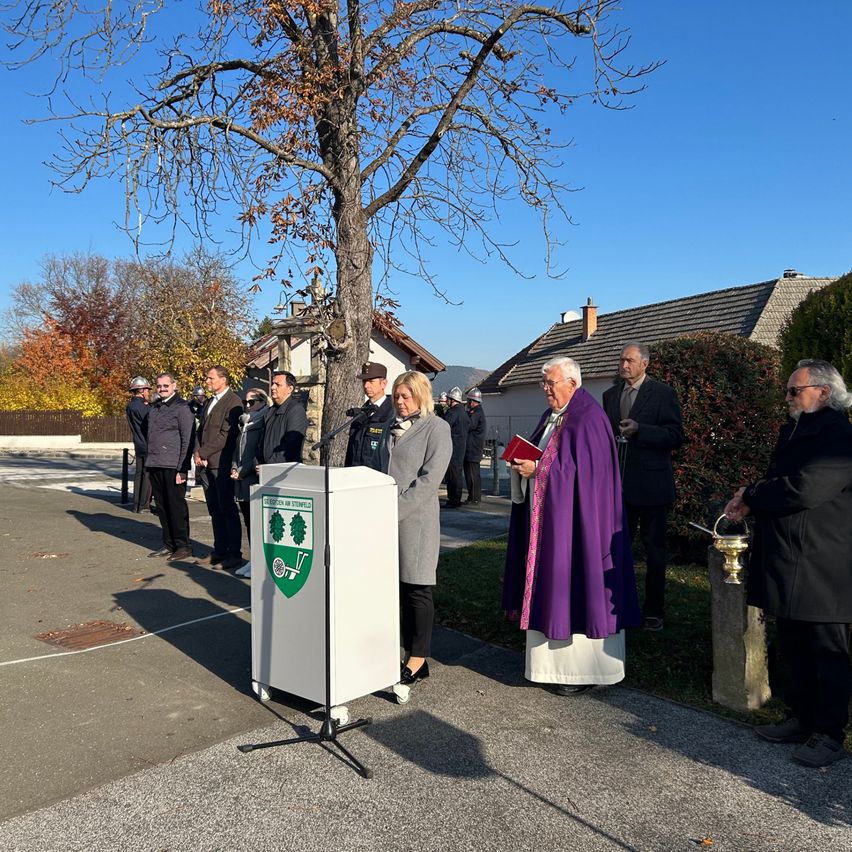 Eine Gruppe von Menschen versammelt sich in einem Park unter einem Baum. Ein Priester steht an einem Rednerpult mit einem Buch und ein Mann hält eine Fackel. Sie sind formell gekleidet.