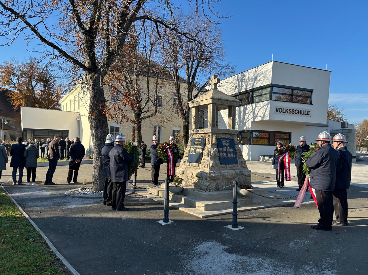 Men in Uniformen stehen um ein Denkmal herum und legen Kränze nieder. Dahinter steht ein Gebäude mit dem Schild 'VOLKSSCHULE'. Das Denkmal hat Tafeln und ein Kreuz oben. Im Hintergrund sind Bäume und ein klarer Himmel zu sehen.