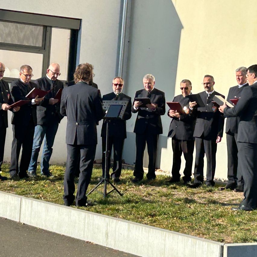 Ein Chor von Männern in schwarzen Anzügen und Brillen steht auf einem Rasenfeld, hält Bücher und singt.