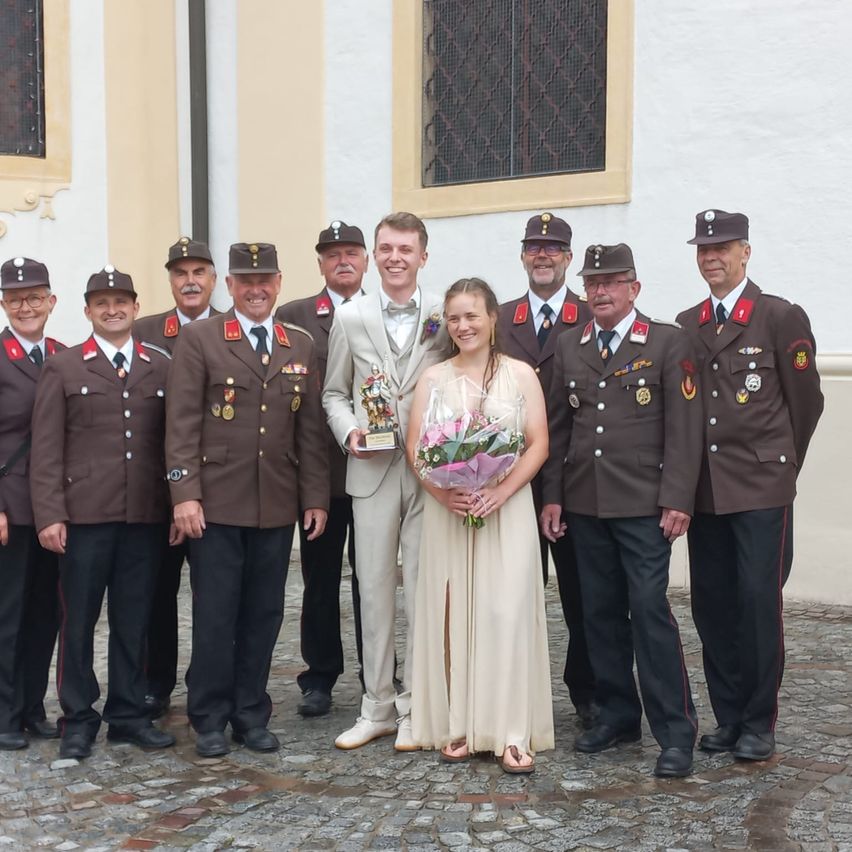 Eine Gruppe uniformierter Männer und eine Frau stehen vor einem Gebäude. Der Mann in der Mitte hält einen Pokal. Die Frau hält einen Blumenstrauß.