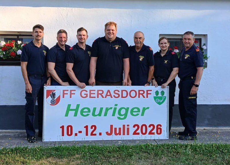 Seven firefighters from FF Gerasdorf pose for a photo in front of a sign. The sign says 'FF Gerasdorf Heuriger 10-12 July 2026'. They are standing on a concrete surface with green grass. Behind them, there is a white wall with a window with red and white flowers.