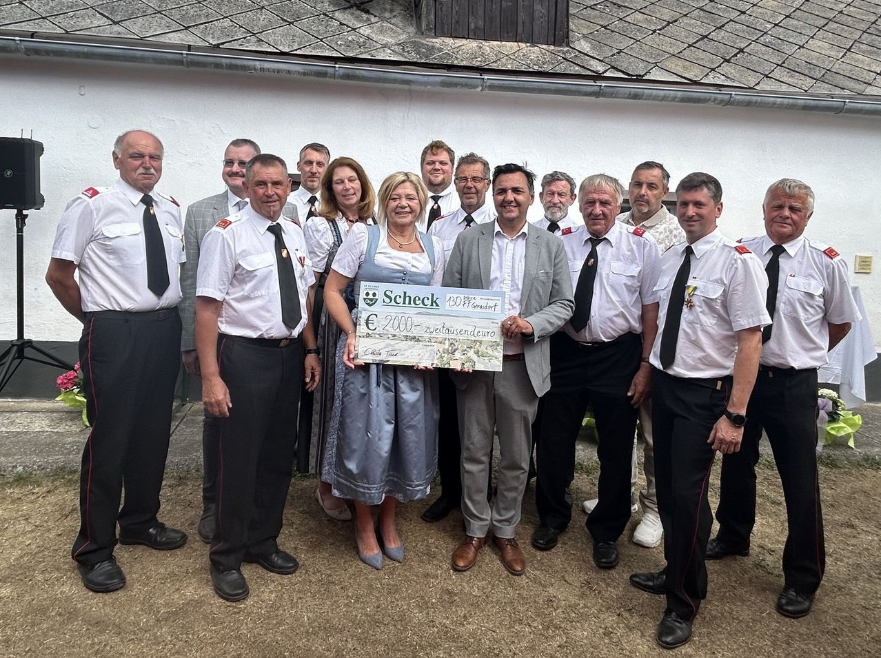 A group of people in uniform, including a woman holding a large check, are standing in front of a building.