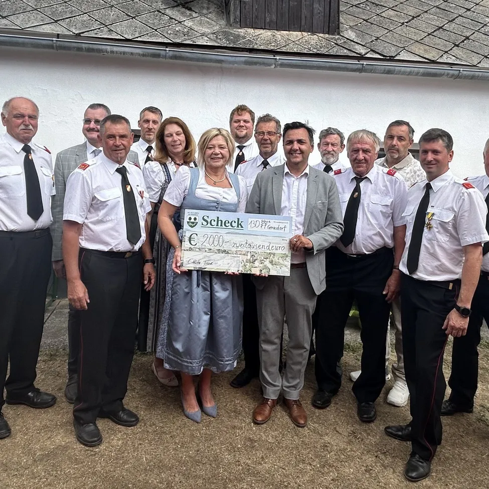 Eine Gruppe von Menschen in Uniform, darunter eine Frau, die einen großen Scheck hält, steht vor einem Gebäude.