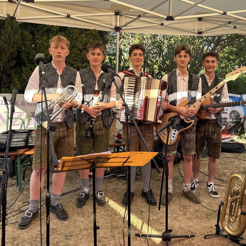 Five young men dressed in traditional Bavarian attire are performing on stage with various musical instruments, including trumpet, accordion, and guitars. They are standing under a white tent.