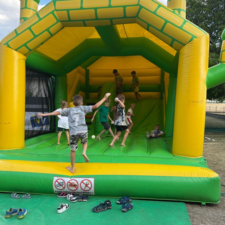 A group of children are playing inside a large inflatable bounce house, some are jumping while others are sitting down. Shoes are placed outside the entrance.