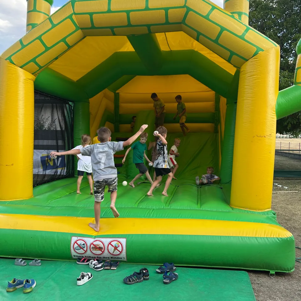 Eine Gruppe von Kindern spielt in einem großen aufblasbaren Hüpfhaus, einige springen, während andere sitzen. Schuhe sind vor dem Eingang platziert.
