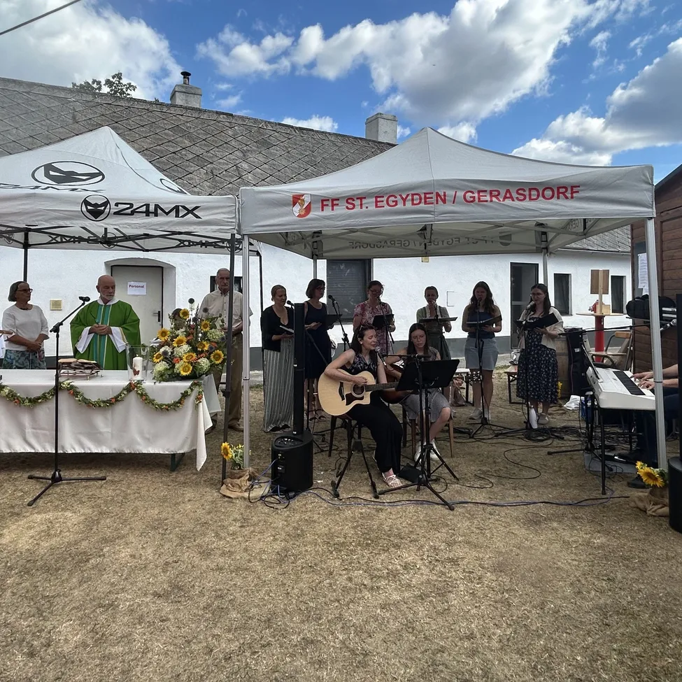Eine Gruppe von Menschen führt ein musikalisches Event unter einem Zelt mit einem Gebäude im Hintergrund auf. Eine Person spielt Gitarre, während andere singen.