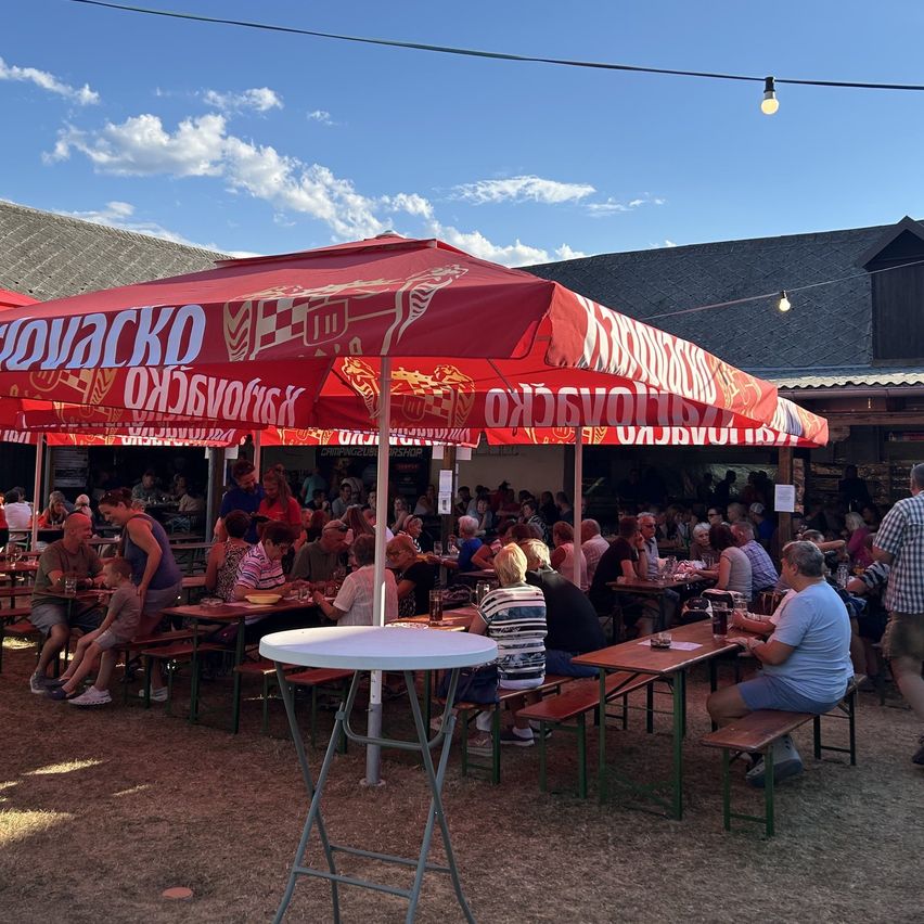 A bustling outdoor dining area with red umbrellas and tables, filled with people enjoying a sunny day. The sky is blue with scattered clouds.