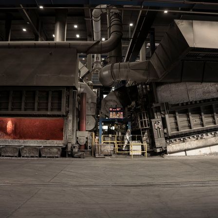 Eine industrielle Fabrik mit einem großen Ofen, Rohren und Maschinen. Eine Person in einer Schutzkleidung steht neben einer gelben Barriere.
