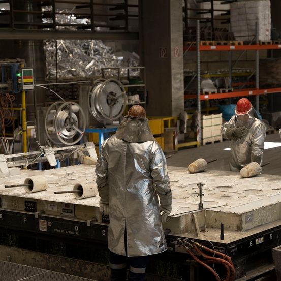 Zwei Arbeiter in Schutzkleidung befinden sich in einer Fabrik. Sie stehen auf einer großen, flachen Plattform. Die Plattform hat Rohre und Schläuche. Die Arbeiter tragen Helme. Im Hintergrund befindet sich ein großer Haufen Altmetall und ein Lagerregal.