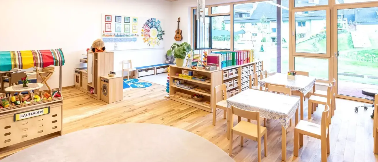 Ein Klassenzimmer mit Holzböden, Tischen und Stühlen. Ein Bücherregal hält Bücher und Ordner. Eine Pflanze und eine Gitarre stehen am Fenster.