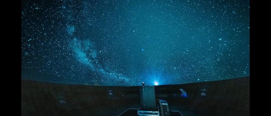 Ein Nachthimmel voller Sterne und einer Galaxie, betrachtet aus einem kuppelförmigen Observatorium. Ausrüstung ist auf dem Boden sichtbar.
