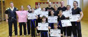 Eine Gruppe von Menschen in Kampfsportuniformen posiert für ein Foto mit Zertifikaten in einer Turnhalle. Einige tragen Gürtel, und ein Basketballkorb ist im Hintergrund.