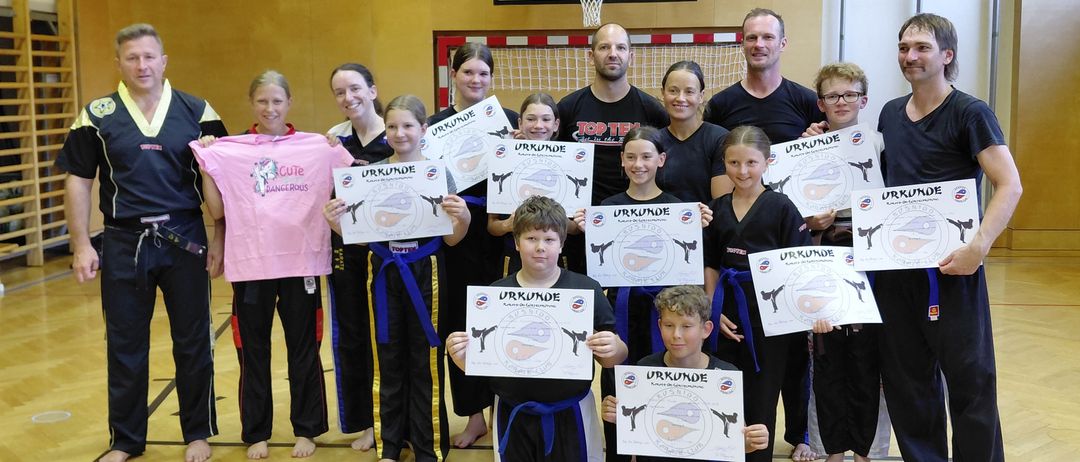 Eine Gruppe von Menschen in Kampfsportuniformen posiert für ein Foto mit Zertifikaten in einer Turnhalle. Einige tragen Gürtel, und ein Basketballkorb ist im Hintergrund.