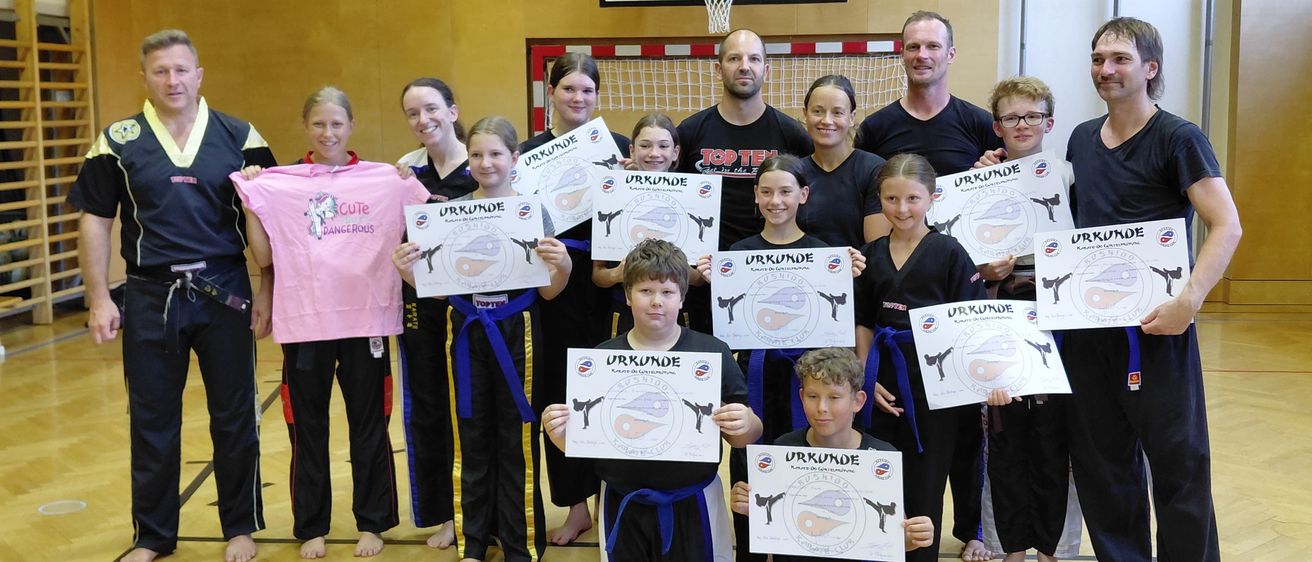 Eine Gruppe von Menschen in Kampfsportuniformen posiert für ein Foto mit Zertifikaten in einer Turnhalle. Einige tragen Gürtel, und ein Basketballkorb ist im Hintergrund.